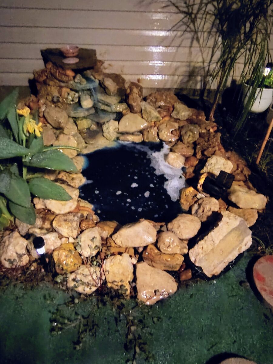 Handmade backyard pond built from stone and reclaimed materials with a small waterfall, lit at night